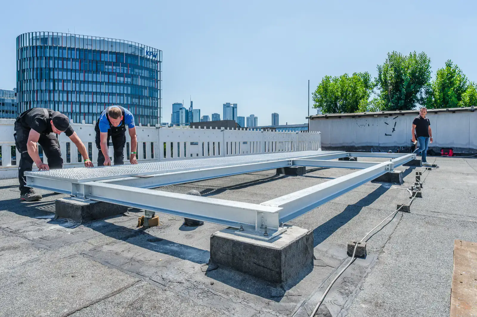 Zwei Monteure richten auf einem Frankfurter Flachdach einen Aluminium-Rahmen aus, im Hintergrund die Skyline